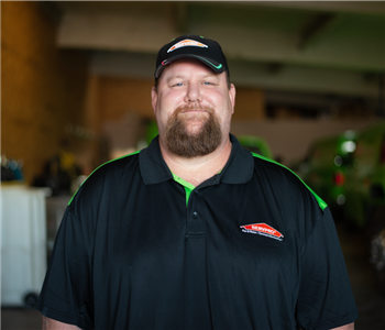 Smiling male employee in a garage wearing a black SERVPRO shirt and black a SERVPRO hat