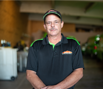 Male employee smiling in a garage wearing a black SERVPRO shirt, and a black SERVPRO hat